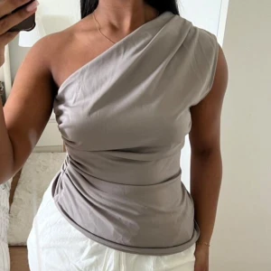 Grå oneshoulder topp - Snygg grå oneshoulder topp med draperad design. Perfekt för en stilren look. Aldrig använd, storlek S, normal i storleken.