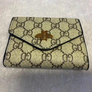 Trendigt Dam  plånbok - Säljer en stilren plånbok med GD-mönster och en dekorativ bi-detalj på framsidan. Plånboken har flera kortfack och en transparent sektion för ID-kort. Finns i två färger: beige och blå.  Helt ny, 89kr/st 