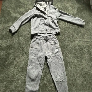 Grå hoodie och byxor från Nike - Säljer en grå hoodie och matchande byxor från Nike. Hoodien har dragkedja och långa ärmar, perfekt för en avslappnad stil. Byxorna har elastisk midja och sidofickor. Båda delarna är i standard fit och tillverkade i mjukt material. Inga skador eller hål, knappt använt. De e barnstorlek