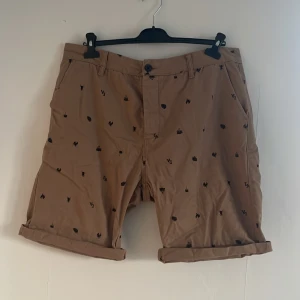 Beige shorts med tryck från Divided - Snygga beige shorts från Divided med ett unikt tryck av små svarta insekter. De har en klassisk passform med knapp och dragkedja framtill. Perfekta för en avslappnad stil med en touch av humor. Märket San Angeles syns på baksidan.