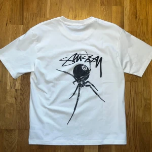 Vit t-shirt med spindeltryck från Stüssy - Snygg vit t-shirt från Stüssy med ett coolt spindeltryck på ryggen och ett mindre tryck på bröstet. Perfekt för en avslappnad stil. Gjord i mjukt material för extra komfort.