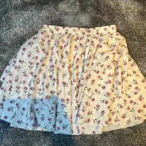 Blommig Kjol - Söt vit kortkjol med blommigt mönster i rosa/lila och grönt. Kjolen har en stretchig resårmidja och den har två lager av tyg för en ”flowig look”! Tyvärr aldrig kommit till användning så den är som ny!
