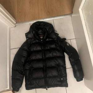 Moncler maya - Säljer en snygg svart pufferjacka från Moncler. Jackan har en quiltad design med långärmade ärmar och en praktisk dragkedja framtill. Perfekt för kyliga dagar med sin varma och stilrena look. Moncler-logga på ärmen ger en exklusiv touch.