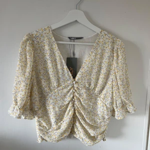 Blommig blus från ONLY - Säljer en söt blommig blus från ONLY i vitt och gult. Blusen har en v-ringad design med knytning framtill och trekvartsärmar. Perfekt för vår och sommar! Aldrig använd, har prislappen kvar. Köptes ny för 350kr. 