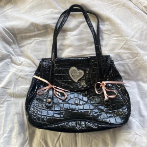 Svart axelväska från Guess - Snygg svart axelväska från Guess i krokodilpräglat syntetmaterial. Väskan har två handtag och dekorativa rosa rosetter på framsidan. Insidan är rymlig med ett mönstrat foder och en dragkedjeficka. Perfekt för att bära dina dagliga nödvändigheter med stil. Osäker på om den äkta då den är köpt här på plick, därav ett lägre pris 