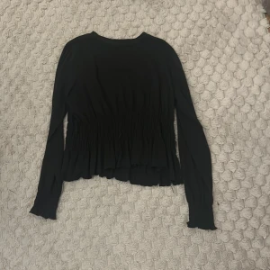 Svart långärmad topp från Vila - Säljer en elegant svart långärmad topp från Vila. Toppen har en smickrande passform med rynkade detaljer i midjan och en vågig nederkant. Perfekt för en stilren look.