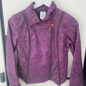 Jacka  Disney Store - Samlarobjekt från Disney Store. och filmen Descendants. Cool lila jacka från Disney Store med dragkedja och nitar. Den har ett unikt broderat mönster på ryggen och en röd äppeldetalj på dragkedjan. Perfekt för att sticka ut med en edgy stil!
