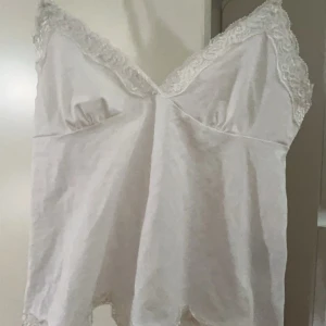 Vit linne med spetsdetaljer - Säljer en söt vit linne med spetsdetaljer längs kanterna. Toppen har en v-ringad design. Den är tillverkad i ett mjukt och bekvämt material.Meddela mig om det är några minsta funderingar💕