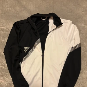 Svart och vit jacka från Adidas - Snygg svart och vit jacka från Adidas med dragkedja och huva. Jackan har ett sportigt utseende med Adidas-loggan på bröstet och en kontrasterande design. Perfekt för en aktiv livsstil.