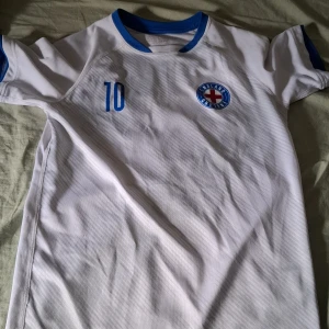 Vit fotbollströja med blå detaljer - Säljer en vit fotbollströja från H&M med blå detaljer. Tröjan har nummer 10 och texten 'England' på ryggen. Den har korta ärmar och en rund halsringning med blå kant. Perfekt för fotbollsfans!