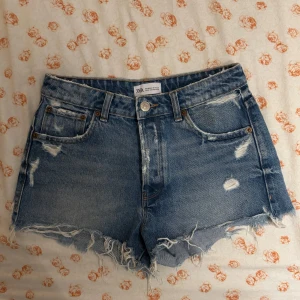 Blå jeansshorts från Zara - Säljer ett par snygga blå jeansshorts från Zara med slitna detaljer och fransig kant. Perfekta för en avslappnad stil. De har en klassisk femficksdesign och knappgylf. 