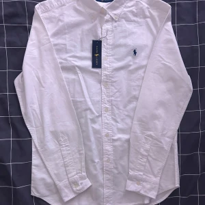 Vit skjorta från Ralph Lauren - Klassisk vit skjorta från Ralph Lauren med broderad logga på bröstet. Skjortan har långa ärmar och knappar framtill. Perfekt för en stilren look.