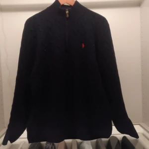 Marinblå stickad tröja från Ralph Lauren - Säljer en snygg mörkblå stickad tröja från Ralph Lauren med dragkedja och det klassiska röda logotypen på bröstet. Nypris sek2795,00. Perfekt för kyliga dagar med sin kabelstickade design och höga krage.