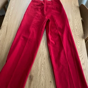 Röda kostymbyxor från MNG - Snygga röda kostymbyxor från Mango med hög midja och raka/vida ben. Perfekta för en stilren look. Byxorna har en klassisk design med diskreta fickor baktill. Finns enstaka trådar som gått upp men påverkar inte helheten.