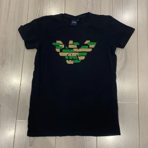 Mörkblå t-shirt från Armani Jeans - Snygg mörkblå t-shirt från Armani Jeans med ett coolt kamouflagemönster i grönt och beige på bröstet. Perfekt för en stilren look. Kortärmad och tillverkad i mjukt material för extra komfort.