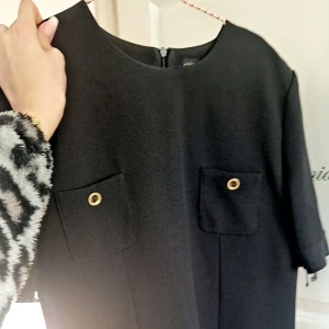 Svart klänning med knappar - Elegant svart klänning med korta ärmar och två dekorativa fickor framtill med knappar. Perfekt för en stilren look.