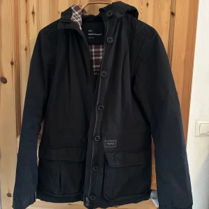 Svart jacka från Peak Performance - Säljer en svart jacka från Peak Performance med huva och knäppning framtill. Jackan har ett rutigt foder i brunt och rött, samt praktiska fickor. Perfekt för kyligare dagar.