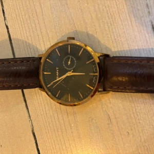 Elegant herrklocka från Gant - Snygg herrklocka från Gant med en stilren design. Klockan har en mörk urtavla med guldiga visare och markeringar. Boetten är i guldton och armbandet är gjort av brunt skinn. Perfekt för den som vill ha en klassisk och tidlös accessoar. Använd, några synliga repor på glaset. Brunt läderarmband i fint skick. Oklart vilken modell det är. Observera att klockan stannat, borde fungera efter ett batteribyte.