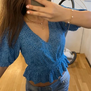 Blå blommig omlottopp - Säljer en söt blå omlottopp med små vita blommor. Toppen har korta ärmar och en volangdetalj vid midjan. Perfekt för en somrig look! 💙