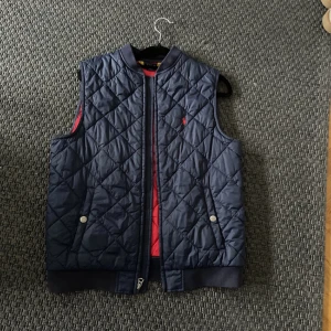 Polo Ralph lauren väst - Snygg mörkblå quiltad väst med dragkedja och knappar. Perfekt för lager-på-lager under kyligare dagar. Den har en diskret broderad logga på bröstet och ribbstickad kant vid ärmslut och nederkant. 9/10 skick.