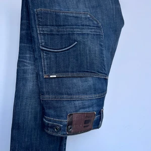G-star jeans - Snygga mörkblå jeansbyxor med unika fickdetaljer och knappar. Perfekta för en cool och avslappnad stil. Märkesdetalj på fickan ger en extra touch.
