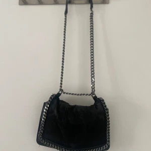 Svart axelväska med kedjedetaljer - Snygg svart axelväska med kedjedetaljer och en unik textur på framsidan. Väskan har en stilren design med en kedja som axelrem, vilket ger en cool look. Perfekt för att ge din outfit en edgy touch.