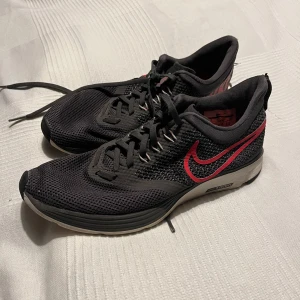 Svarta Nike löparskor  - Snygga svarta Nike löparskor med röd swoosh och snörning. Skorna har en bekväm sula med bra grepp och är perfekta för löpning eller träning. Ovandelen är i ett andningsbart material för extra komfort.
