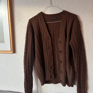 Brun stickad kofta från Brandy Melville - Mysig brun stickad kofta från BrandyMelville med kabelstickat mönster. Koftan har långa ärmar och knappar framtill, perfekt för kyliga dagar. En klassisk och tidlös design som passar till det mesta.