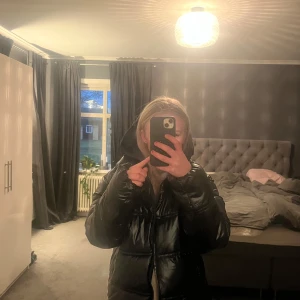 Svart Massimo dutti jacka - Säljer en svart pufferjacka med huva och dragkedja. Jackan är långärmad och perfekt för kyligare väder. Den har en glansig yta som ger en modern look. Liten i storlek!