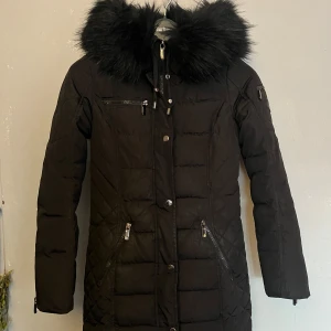 Svart dunjacka med pälsdetaljer - Snygg svart dunjacka från Marikoo med quiltad design och pälsdetaljer på huvan. Jackan har dragkedja och knappar framtill samt flera praktiska fickor. Insidan är fodrad med ett leopardmönster som ger en cool kontrast. Perfekt för kalla vinterdagar. Jackan är något längre och går över rumpan. Defekter förekommer: det är hål i båda armhålorna på insidan ( se bild 3 ), samt en missfärgning på framsidan utav jackan ( se bild 4 ). 