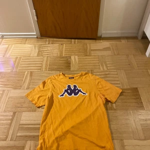 Orange t-shirt från Kappa - Säljer en cool orange t-shirt från Kappa med deras ikoniska logga i vitt och svart på bröstet. Perfekt för en avslappnad stil. Tröjan är storlek M