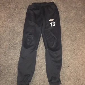 Svarta träningsbyxor från Umbro - Säljer ett par svarta träningsbyxor från Umbro med elastisk midja och nummer 13 tryckt på framsidan. Byxorna har vita detaljer längs benen och är perfekta för sportaktiviteter.