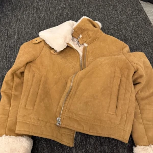 Beige mockajacka med foder - Säljer en snygg beige mockajacka med mjukt foder och dragkedja framtill. Jackan har långa ärmar och en hög krage som ger extra värme. Perfekt för kyliga dagar!