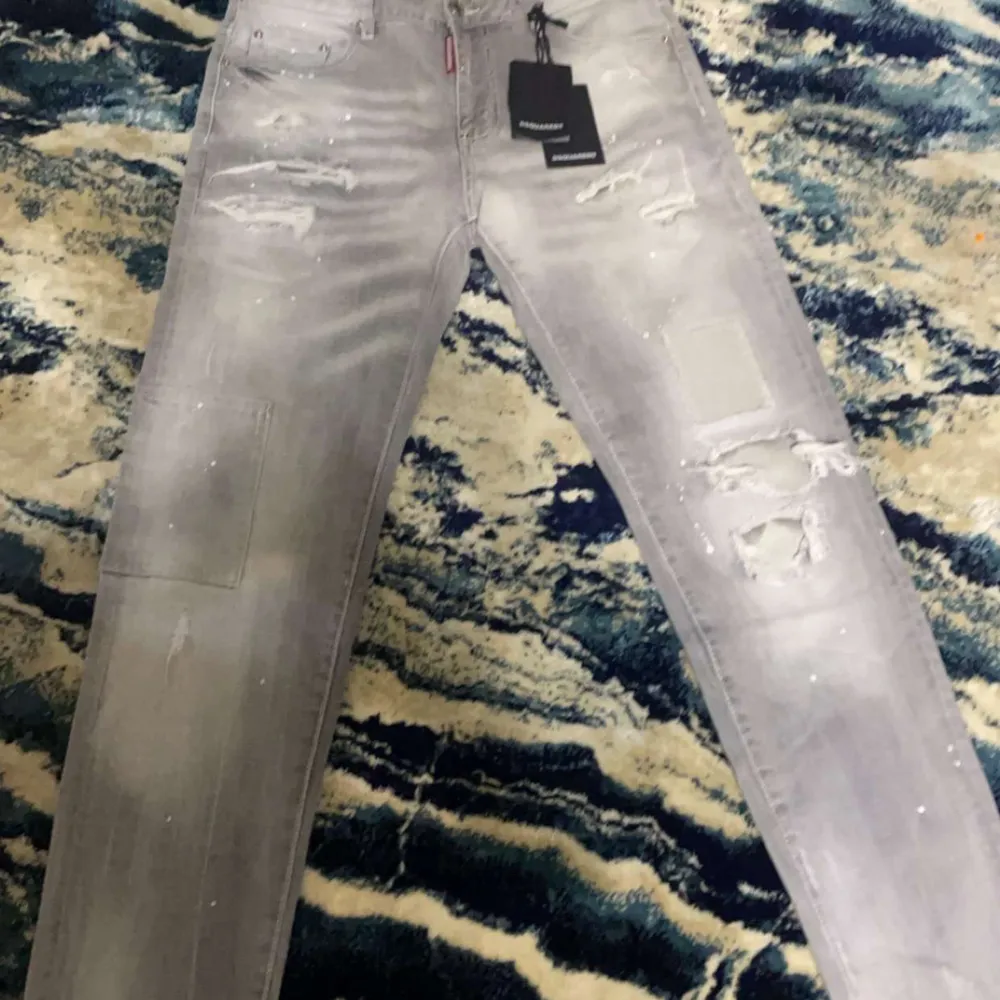 Säljer ett par snygga grå jeans från Dsquared2 i nytt skick. De har en cool distressed look med slitningar och färgstänk. Perfekta för en trendig och avslappnad stil. Tillverkade i Italien med hög kvalitet.. Jeans & Byxor.