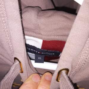 Säljer en snygg beige hoodie från Tommy Hilfiger i storlek XS. Den har en stor logga broderad på framsidan och en praktisk magficka. Perfekt för en avslappnad stil och passar bra till både jeans och joggers. Huvan har justerbara snören för extra komfort. Använd 3 gånger 