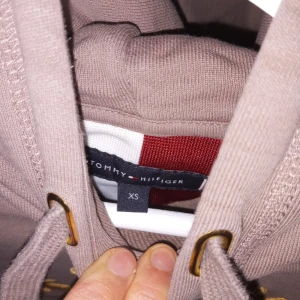 Beige hoodie från Tommy Hilfiger - Säljer en snygg beige hoodie från Tommy Hilfiger i storlek XS. Den har en stor logga broderad på framsidan och en praktisk magficka. Perfekt för en avslappnad stil och passar bra till både jeans och joggers. Huvan har justerbara snören för extra komfort. Använd 3 gånger 