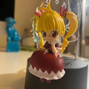 Hunter x hunter figur - En supergullig animefigur med blont hår i två tofsar och en röd klänning med vita detaljer. Hon sitter på en stenliknande bas och håller en stjärnformad stav. Perfekt för samlare eller som dekoration på skrivbordet!