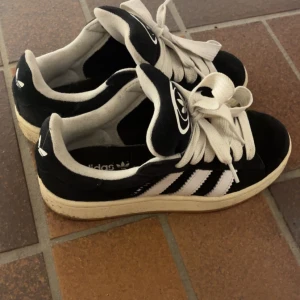 Svarta Adidas Campus 00 - Snygga svarta Adidas campus i storlek  36 2/3 i men dem sitter som en 37  sneakers med vita detaljer och snörning. Klassisk design med tre ränder på sidan och en bekväm sula. Perfekta för en avslappnad stil.