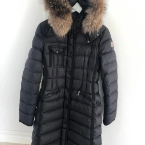 Svart dunjacka med pälsdetalj - Snygg svart dunjacka från Moncler med pälsdetalj på huvan. Jackan har en quiltad design och långa ärmar. Perfekt för kalla vinterdagar med sin varma och stilrena look. Jag har fortfarande det fysiska orginalkvittot från Olséns som följer med. Jackan är i otroligt bra skick! 