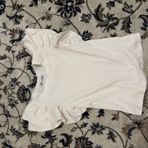 Vit ribbad topp från Zara - Säljer en söt vit ribbad topp från Zara i storlek S. Den har korta ärmar med volangdetaljer som ger en romantisk touch. Perfekt för både vardag och festliga tillfällen. Toppen är i mycket bra skick och redo för en ny ägare!