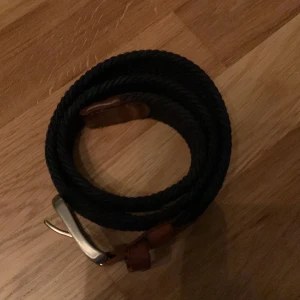 Blått flätat bälte storlek 80 cm - Säljer ett snyggt blått flätat bälte från Jack and Jones med bruna läderdetaljer och en stilren silverfärgad spänne. Perfekt för både vardag och fest! En liten del av  lacken på spänet har got bort som man ser på bild 4