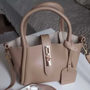 Beige handväska med gulddetaljer - Elegant beige handväska med gulddetaljer och avtagbar axelrem. Väskan har en stilren design med ett dekorativt spänne framtill och en praktisk namntag. Perfekt för att ge en touch av lyx till din outfit.