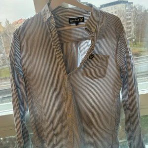 Blå och vit randig skjorta från Lyle & Scott - Säljer en stilren blå och vit randig skjorta från Lyle & Scott i storlek M. Skjortan har långa ärmar och en klassisk krage. Den är tillverkad i bomull och har en liten broderad logotyp på bröstfickan. Perfekt för både vardag och fest!
