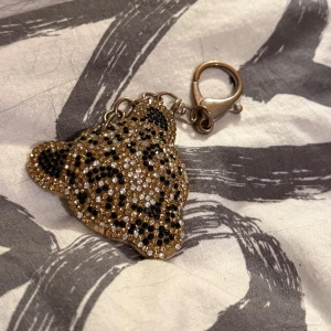 leopard nyckelring - säljer en snygg nyckelring i form av ett leopardhuvud, täckt med glittrande strass i guld, svart och silver. pris kan alltid diskuteras💕(hundhår i bakgrunden)