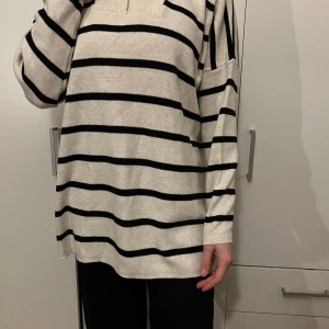 Randig beige och svart tröja - Säljer en supermysig randig tröja i beige och svart. Den har en loose passform och långa ärmar, perfekt för kyliga dagar. Tröjan har en dragkedja vid halsen som ger en cool detalj. Perfekt för både höst och vår!
