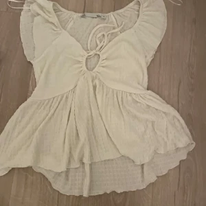 Vit blus med volangdetaljer - Charmig vit blus med volangdetaljer och knytning framtill. Blusen har en luftig passform och korta ärmar, perfekt för varma dagar. Den har en v-ringad design med en liten öppning framtill som ger en extra touch.