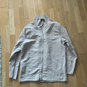 Beige kofta från H&M - Säljer en stilren beige kofta från H&M med dragkedja framtill. Perfekt för lager-på-lager under kyligare dagar. Koftan har en enkel design med långa ärmar och en hög krage.