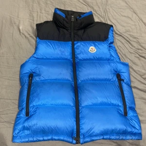 Moncler Väst - Säljer en snygg blå och svart dunväst från Moncler. Västen har en dragkedja framtill och två praktiska fickor med dragkedjor. Perfekt för kyliga dagar och ger en stilren look. Moncler-loggan är broderad på bröstet. Passar både för höst och vår. Köpt för 10k i Nk