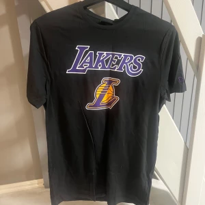 Svart Lakers t-shirt från New Era - Snygg svart t-shirt från New Era med Lakers-logga i lila och gult på bröstet. Perfekt för basketfans som vill visa sitt stöd. T-shirten har en klassisk passform och korta ärmar.