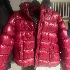 Röd pufferjacka med huva - Snygg röd pufferjacka med huva och dragkedja. Jackan har en glansig finish och är perfekt för vintern nu. Tyvärr kommer den inte till användning längre men den är som ny. Den är i storlek 44 men de är väldigt små i stoleken så jag som brukar ha S eller M på jackor kan ha den här storleken. 